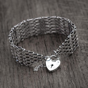 Könnte beinhalten: Ein silberfarbenes Armband mit gewebtem Design. Das Armband hat einen herzförmigen Vorhängeschloss-Anhänger. Das Armband besteht aus mehreren Reihen miteinander verbundener Glieder.