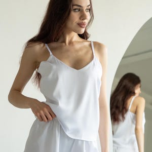 Op de afbeelding: Witte satijnen pyjamaset. De set bestaat uit een topje met dunne bandjes en een V-hals, gecombineerd met bijpassende shorts. Het model draagt de set voor een spiegel.