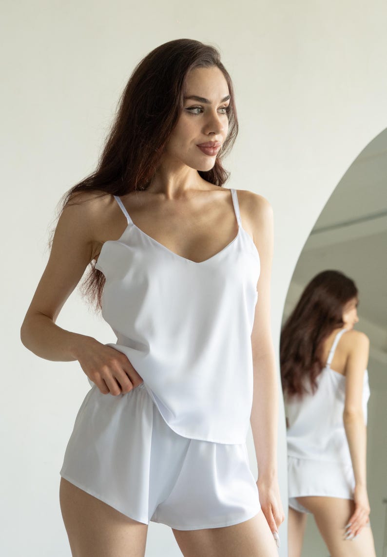 Op de afbeelding: Witte satijnen pyjamaset. De set bestaat uit een topje met dunne bandjes en een V-hals, gecombineerd met bijpassende shorts. Het model draagt de set voor een spiegel.