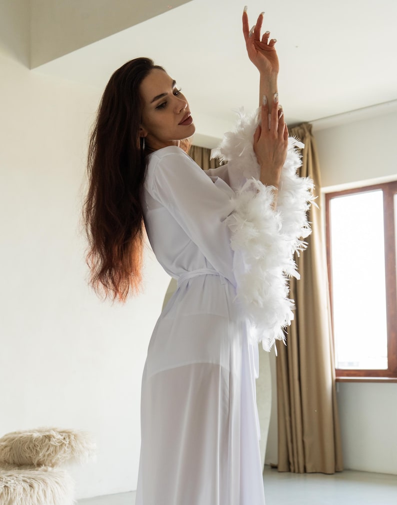 Op de afbeelding: Een witte, doorschijnende badjas met veren op de mouwen. De badjas wordt in de taille vastgemaakt met een bijpassende riem. Het model draagt de badjas in een lichte kamer.