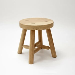 May include: A small, round wooden stool with a light brown finish. The stool has a circular seat and three angled legs connected by horizontal supports. The stool is approximately 30 cm tall.