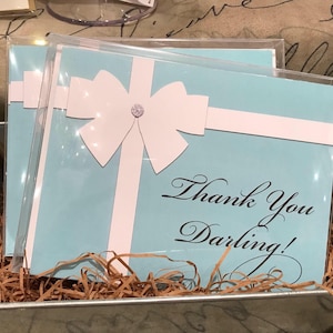 May include: Light blue greeting cards with white ribbon and gem embellishments, reading "Thank You Darling!" in elegant script. The cards are in clear plastic sleeves, nestled in a metal container with brown paper filler.
