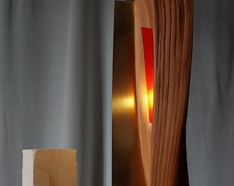 May include: A contemporary wooden sculpture featuring a brass element and a red illuminated section. The sculpture is mounted on a stone base. A second, smaller, white stone sculpture is to the left.