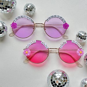 May include: Two pairs of round sunglasses with gold frames and gradient lenses. One pair has purple lenses, the other pink. Both have beaded text that reads "TOGETHER" and "ALL THE TIME". Each pair has a flower accent.