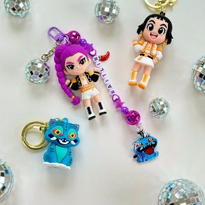 Może przedstawiać: Kolekcja breloków z postaciami z kreskówek i niebieskim kotem. Jeden brelok ma postać z fioletowymi włosami z imieniem "KAITLIN" na zawieszce. Druga postać ma na sobie żółto-biały strój. Rozrzucone są akcenty w postaci kul dyskotekowych.