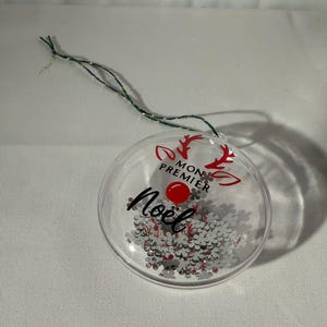 Puede incluir: Adorno navideño transparente con confeti de copos de nieve plateados y las palabras "Mon Premier Noël" en escritura negra. El adorno tiene un punto rojo y cuernos de reno. Una cuerda verde y plateada está adjunta.