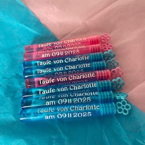 Op de afbeelding: Een stapel heldere, cilindrische gunsten met bloemvormige toppers. De gunsten zijn in roze en blauwe tinten, met de tekst "Taufe von Charlotte am 09 11 2025". De achtergrond is roze en blauw vloeipapier.