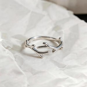 May include: A silver-colored ring with a branch-like design. The ring features a delicate, openwork pattern with multiple small, curved branches. The ring is set against a white, textured background.