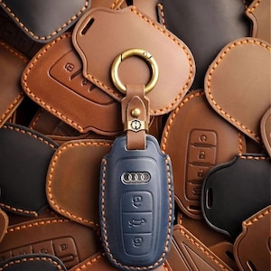 May include: A blue leather car key fob cover with an Audi logo and key buttons, attached to a brown leather strap and gold-colored ring. The fob is surrounded by various brown and black leather key fob covers.