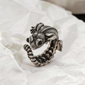 May include: A silver-toned ring featuring a detailed lizard design. The lizard has textured skin, small hands, and black eyes. The ring band has a textured, ribbed design. The ring is set against a white, textured background.