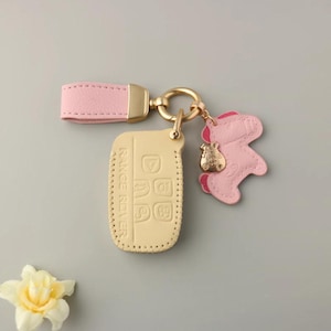 May include: A light pink leather key fob holder with a gold-tone metal clasp and a matching pink leather strap. The fob holder is for a Range Rover key and has a small pink bear charm.