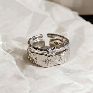 Può includere: Set di anelli in argento con fascia strutturata e design a stella. L'anello superiore presenta una gemma a forma di stella. L'anello è contrassegnato con "S925". L'anello è su una superficie bianca strutturata.