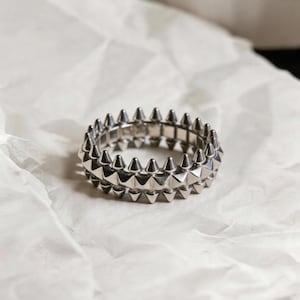 May include: A silver-colored ring with a double-band design. Each band is adorned with evenly spaced, pointed spikes. The ring is displayed on a white, textured surface, creating a contrast with the metallic sheen of the jewelry.