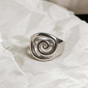May include: A silver ring with a spiral design. The ring is made of a shiny metal and has a thick band. The spiral is centered on the ring's face and is open, allowing light to pass through.
