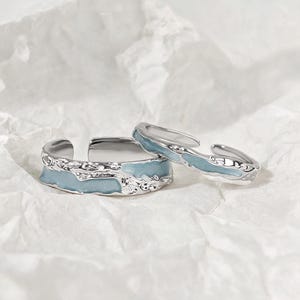 May include: Two open-band rings featuring a silver-toned metallic finish with a textured, irregular surface. Each ring is accented with a smooth, light blue enamel inlay that follows the organic, wavy contours of the metal band design.