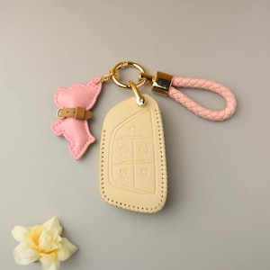 Puede incluir: Un juego de llavero y funda para llave. La funda para llave es de color beige claro con iconos de botones en relieve. El llavero incluye un dije de oso de cuero rosa y una correa de muñeca trenzada rosa, todo conectado por herrajes dorados.