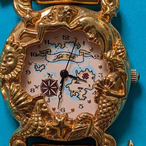 May include: A gold-tone wristwatch with a decorative bezel featuring sculpted mermaids, a sun, and shells. The watch face displays a map illustration with the text Kinks Folly, a compass rose, and black hands. The band includes star-shaped charms.