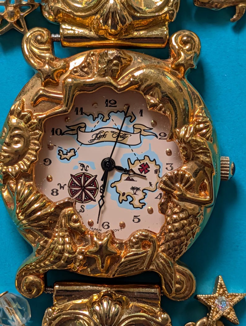 May include: A gold-tone wristwatch with a decorative bezel featuring sculpted mermaids, a sun, and shells. The watch face displays a map illustration with the text Kinks Folly, a compass rose, and black hands. The band includes star-shaped charms.