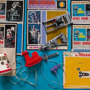 May include: A collection of vintage Mattel's Man in Space toy sets. The image shows several boxes and accessories, including a red and white space capsule, a red vehicle, and a gray launch platform. The boxes feature illustrations of astronauts.