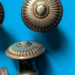 May include: Close-up of several antique brass cabinet knobs. The knobs have a circular design with a central button and radiating lines. The knobs are a warm gold color against a blue background.