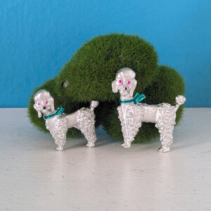 May include: Two silver poodle brooches with pink eyes and teal bows around their necks. The brooches are detailed with textured fur and stand on a white surface against a green mossy background and a blue wall.