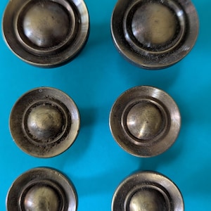 May include: Six circular cabinet knobs made of antique brass metal. Each knob features a central raised dome surrounded by a flat, wide circular rim. The metal has a weathered, dark gold finish with visible surface texture and slight variations.