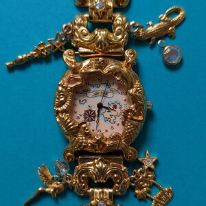 May include: A gold-tone analog wristwatch featuring a map-themed dial with a compass rose and landmass illustrations. The bezel is decorated with embossed sun, mermaid, and shell motifs. The bracelet includes charms of a star, alligator, fairy, and palm tree.