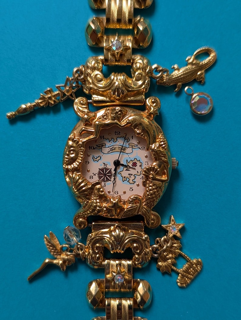 May include: A gold-tone analog wristwatch featuring a map-themed dial with a compass rose and landmass illustrations. The bezel is decorated with embossed sun, mermaid, and shell motifs. The bracelet includes charms of a star, alligator, fairy, and palm tree.