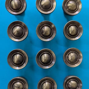May include: A set of twelve circular cabinet knobs arranged in a grid. Each knob features a metallic bronze finish with a raised central dome surrounded by a recessed ring and a wide, flat outer rim, creating a uniform and functional hardware design.