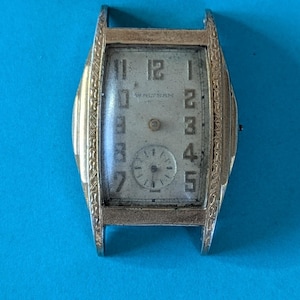 May include: Vintage rectangular wristwatch with a silver-tone case and a light-colored face. The watch features black numerals, a small seconds dial, and the brand name "Waltham". The watch is set against a blue background.