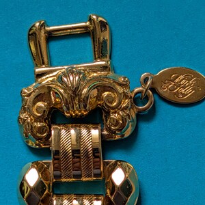 May include: A gold-tone metal bracelet featuring a decorative buckle-style clasp with ornate scrollwork. The band consists of alternating polished diamond-shaped links and textured rectangular links with diagonal stripes. An oval tag reads Kink A Folly.