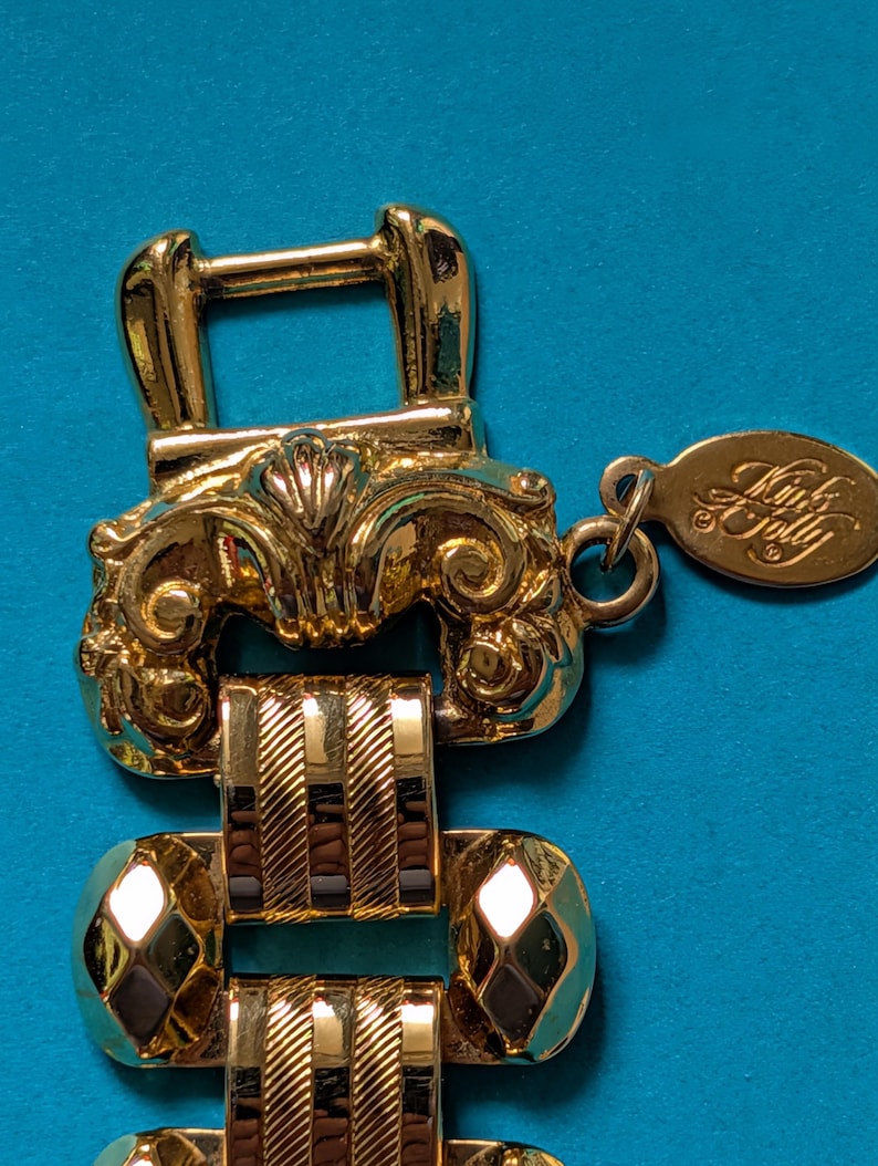 May include: A gold-tone metal bracelet featuring a decorative buckle-style clasp with ornate scrollwork. The band consists of alternating polished diamond-shaped links and textured rectangular links with diagonal stripes. An oval tag reads Kink A Folly.