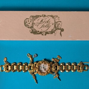 May include: A gold-tone link watch with a round face and multiple dangling charms, featuring an ornate frame and a light-colored dial. The watch is accompanied by a rectangular light pink box with the text Kirks Folly printed in dark brown.