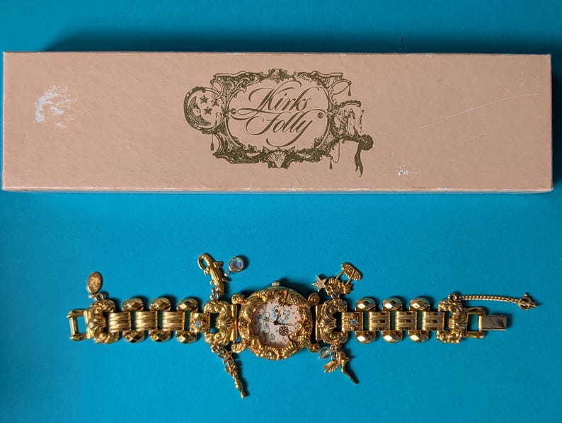 May include: A gold-tone link watch with a round face and multiple dangling charms, featuring an ornate frame and a light-colored dial. The watch is accompanied by a rectangular light pink box with the text Kirks Folly printed in dark brown.
