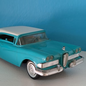 May include: A turquoise and white scale model car featuring a prominent vertical chrome grille, dual round headlights on each side, a silver front bumper, and white-walled tires. The vehicle has a sleek body design with a white roof and clear windows.