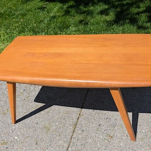 May include: A mid-century modern style coffee table with a rectangular top and angled legs. The table is made of light-colored wood, likely teak or a similar material. The table measures approximately 48 inches long and 20 inches wide.
