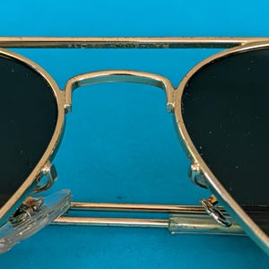 Op de afbeelding: Close-up van een goudkleurige zonnebril met donkere glazen. De brug en de pootjes zijn goudkleurig en de glazen zijn donkergroen. De zonnebril staat tegen een helderblauwe achtergrond.