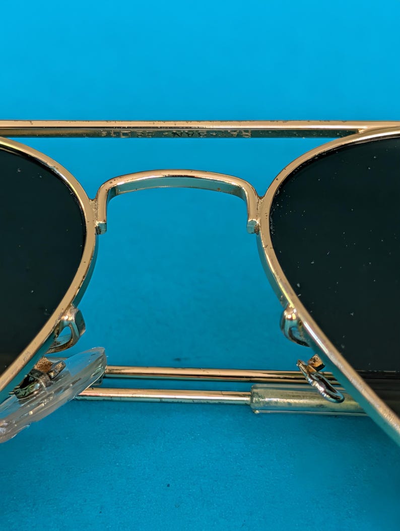 Op de afbeelding: Close-up van een goudkleurige zonnebril met donkere glazen. De brug en de pootjes zijn goudkleurig en de glazen zijn donkergroen. De zonnebril staat tegen een helderblauwe achtergrond.