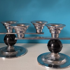 May include: Two silver-toned candelabras with six candle holders each. The holders have a tiered design. Each candelabra has a black sphere in the middle, and a round base. The background is a solid teal color.