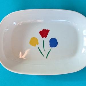 May include: A white, oval-shaped ceramic serving platter with rounded edges. The platter features a colorful floral design with a red, yellow, and blue flower. The flowers have green stems. The background is a solid turquoise color.