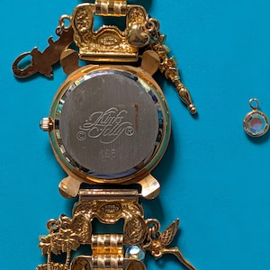 May include: A gold-tone charm watch with a metal link band featuring various dangling charms including palm trees and a fairy. The watch back is engraved with the text Kink's Folly and K35. A separate small circular charm with a clear stone is included.