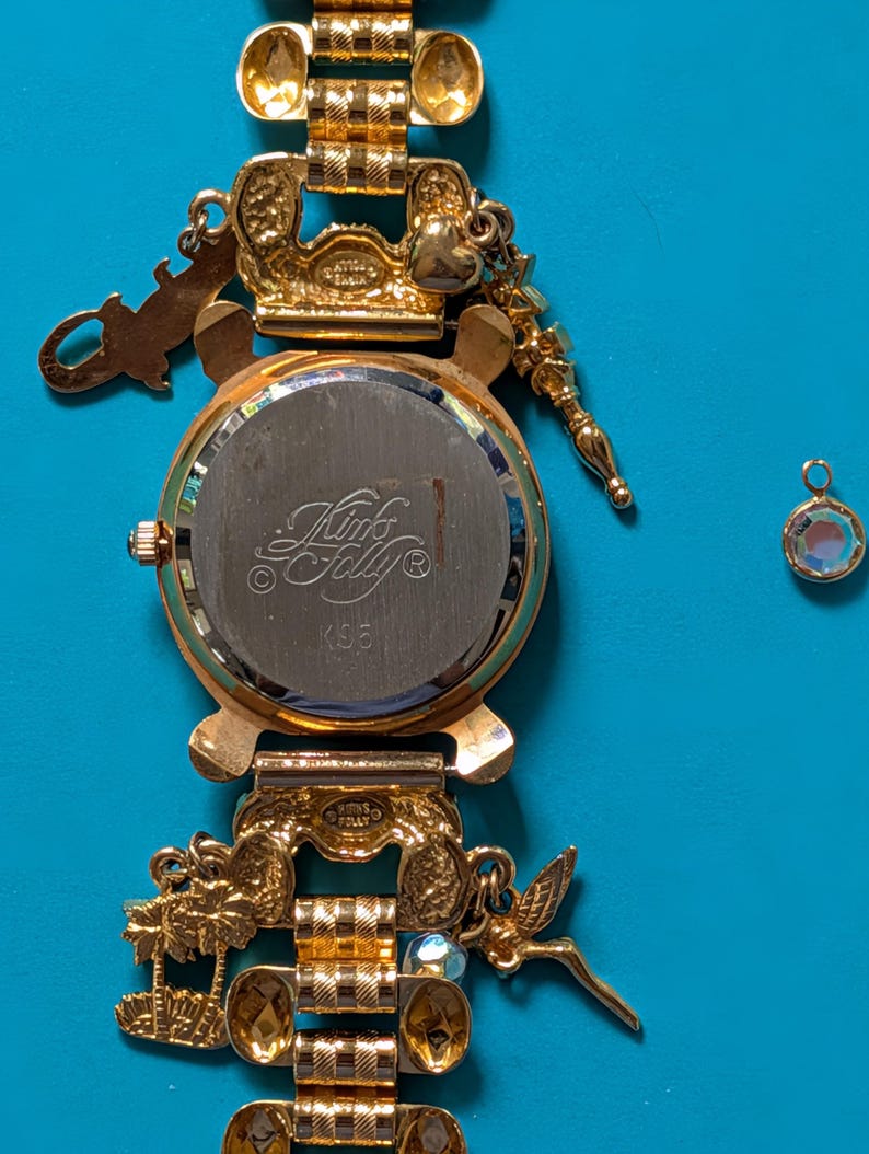 May include: A gold-tone charm watch with a metal link band featuring various dangling charms including palm trees and a fairy. The watch back is engraved with the text Kink's Folly and K35. A separate small circular charm with a clear stone is included.