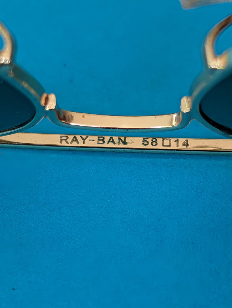 Op de afbeelding: Close-up van een Ray-Ban zonnebril met goudkleurig montuur en donkere glazen. De tekst "RAY-BAN 58 14" staat op het montuur. De achtergrond is effen blauw.