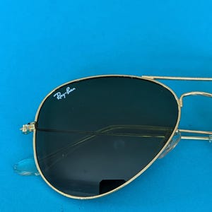 Op de afbeelding: Goudkleurige pilotenbril met donkere glazen. Het Ray-Ban-logo is zichtbaar op het linkerbovenglas. De zonnebril staat tegen een helderblauwe achtergrond.