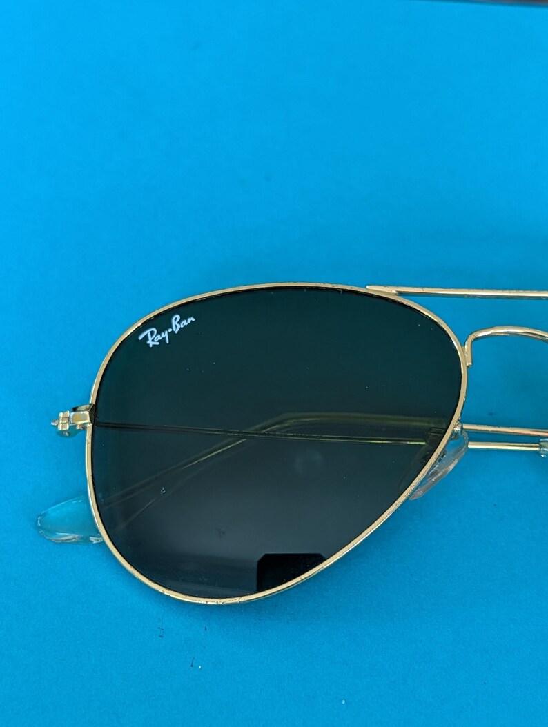 Op de afbeelding: Goudkleurige pilotenbril met donkere glazen. Het Ray-Ban-logo is zichtbaar op het linkerbovenglas. De zonnebril staat tegen een helderblauwe achtergrond.