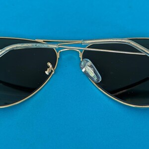 Op de afbeelding: Aviator zonnebril met een goudkleurig metalen montuur en donkere glazen. De zonnebril heeft transparante kunststof oorpootjes en neuspads. De bril ligt op een helderblauwe achtergrond.