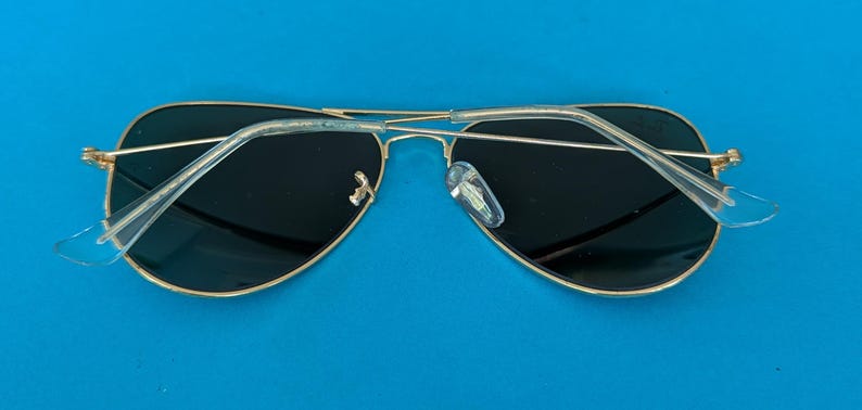 Op de afbeelding: Aviator zonnebril met een goudkleurig metalen montuur en donkere glazen. De zonnebril heeft transparante kunststof oorpootjes en neuspads. De bril ligt op een helderblauwe achtergrond.