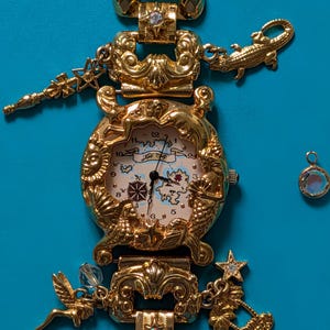 May include: A gold-tone charm watch featuring a map-themed dial with the text 'Foli Follie'. The watch band is adorned with various gold-tone charms including a crocodile, a fairy, a palm tree, and stars, with some charms accented by clear crystals.