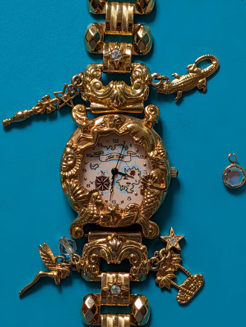 May include: A gold-tone charm watch featuring a map-themed dial with the text 'Foli Follie'. The watch band is adorned with various gold-tone charms including a crocodile, a fairy, a palm tree, and stars, with some charms accented by clear crystals.