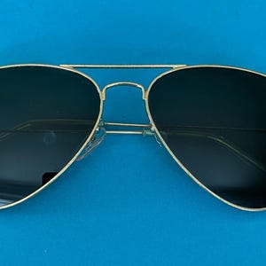 Op de afbeelding: Aviator zonnebril met een goudkleurig montuur en donkere glazen. De zonnebril heeft een dubbele brug en transparante kunststof oortips. Het Ray-Ban logo is zichtbaar op het linkse glas. Op een blauwe achtergrond.